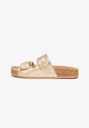 Beige slip-on sandal with cork sole and two large gold buckle straps across the top, flat with textured rubber outsole.