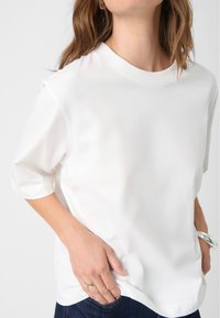 White short-sleeve top with a crew neck, smooth texture, relaxed fit, and side slit details, paired with dark denim jeans.