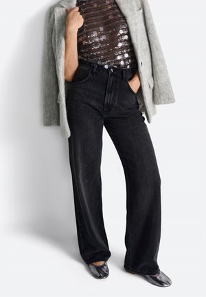 Zwarte high-waisted wijde jeans met een knoopsluiting, gecombineerd met een pailletten top en een lichtgrijs wollen jas. Glanzende zwarte ballerina's maken de look af.
