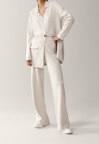Jasno beżowa marynarka dopasowana z jednym guzikiem i kieszenią, zestawiona z szerokimi spodniami w tym samym odcieniu oraz białymi sneakersami. Gładka tekstura.