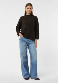 Maglione a coste in marrone scuro con collo alto, abbinato a jeans a gamba larga di un blu chiaro. Il maglione presenta un motivo a treccia centrale.