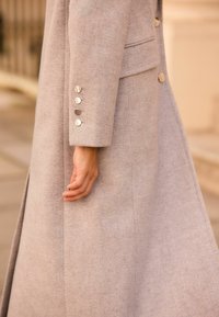 Manteau en laine gris clair avec un tissu texturé, présentant une coupe ajustée, une fente sur le côté et des boutons métalliques au niveau des poignets.