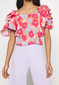 Blumiges Crop-Top in Pink, Orange und Blau mit Rüschen-Ärmeln, strukturiertem Stoff und quadratischem Ausschnitt, kombiniert mit helllila Unterteilen.