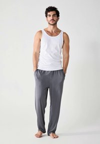 Un débardeur en coton blanc et un pantalon de détente en tissu gris. Les pantalons ont une coupe ample avec une taille élastique et des poches latérales. Position pieds nus.