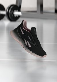 Μαύρο και ροζ αθλητικό παπούτσι Reebok που αιωρείται πάνω από το πάτωμα ενός γυμναστηρίου με μια μπάρα βαρών στο θολό φόντο.