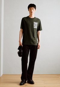 Mørkegrøn bomulds t-shirt med grafisk lomme-design, parret med sorte bukser. Modellen holder en sort taske og står på et trægulv.