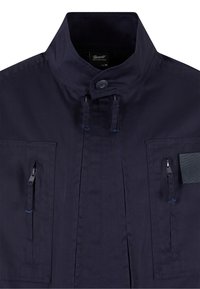 Veste bleu marine foncé avec col montant, fermeture à boutons, deux poches poitrine verticales zippées et patch rectangulaire sur le côté droit.