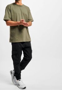 Olijfgroen t-shirt met een ontspannen pasvorm, gecombineerd met zwarte cargobroeken met zakken. Schoenen zijn wit met zwarte accenten en in een hardloopstijl.