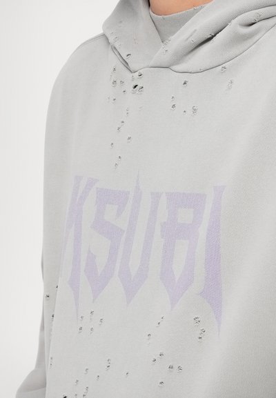 Pilkas džemperis su sugadinta tekstūra ir mažomis skylutėmis, o priekyje yra didelis violetinis logotipas su užrašu "Ksubi".