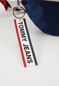 Portachiavi Tommy Jeans con design rettangolare, caratterizzato da bold text nero su sfondo bianco, arricchito da strisce rosse e blu navy.