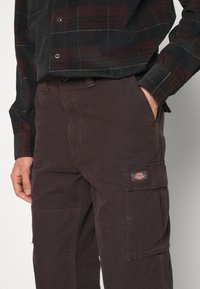 Pantalon cargo marron en tissu robuste, avec poches latérales et patch logo. Associé à une chemise noire à carreaux.
