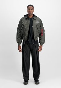 Young man standing wearing an olive green bomber jacket with patches, black hoodie, black wide-leg pants, and black shoes, neutral background.