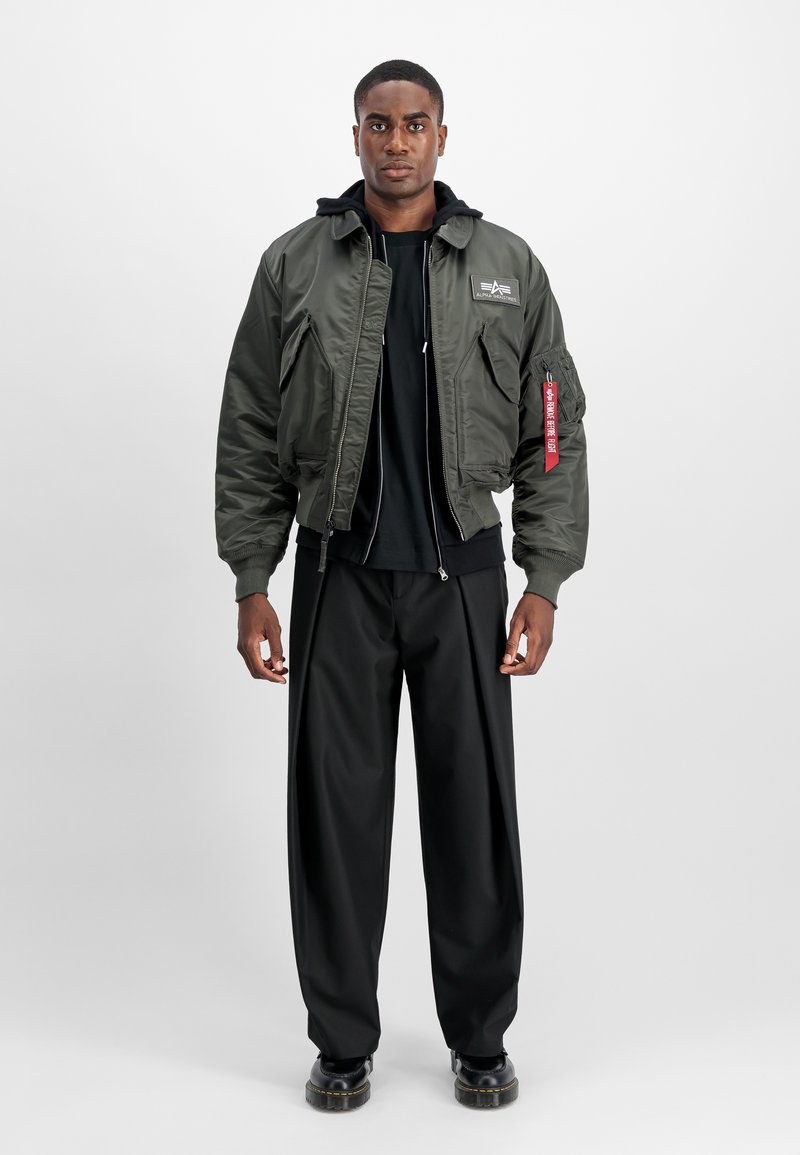Young man standing wearing an olive green bomber jacket with patches, black hoodie, black wide-leg pants, and black shoes, neutral background.
