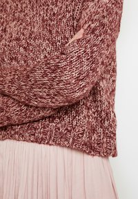 Braccia incrociate in un maglione lavorato a maglia bordeaux e bianco sopra una gonna plissettata rosa chiaro su sfondo bianco.