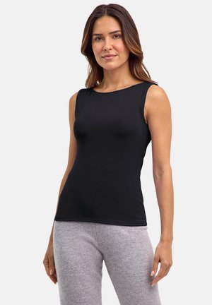 Donna con top nero senza maniche e pantaloni grigio chiaro, in piedi contro uno sfondo grigio chiaro uniforme.