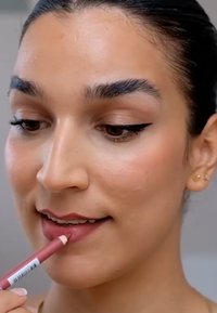 Crayon à lèvres en rose doux appliqué sur les lèvres ; le modèle a une peau lisse, des sourcils bien dessinés, un eyeliner noir et un fond neutre.
