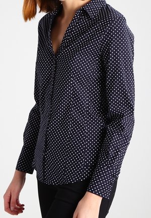 Kobieta w granatowej koszuli z długim rękawem w białe kropki, w czarnych spodniach, na jednolitym tle.