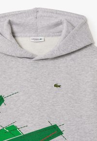 Felpa grigia con logo di coccodrillo verde, caratterizzata da un design grafico con accenti verdi e rossi. Tessuto morbido e texture, vestibilità comoda.