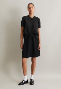 Schwarzes kurzärmeliges Kleid mit Knopfleiste, elastischem Bund und geradem Schnitt. Kombiniert mit schwarzen Schuhen und weißen Socken. Mattes Material.