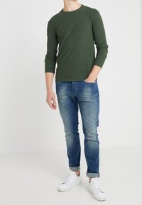 Grön långärmad tröja med rund hals, kombinerad med ljusblå slitna jeans och vita sneakers, vilket visar en avslappnad och bekväm outfit.