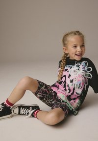 T-shirt noire surdimensionnée avec un graphique floral coloré, associée à un short noir à imprimé floral. Complété par des baskets noires et des chaussettes à rayures rouges.