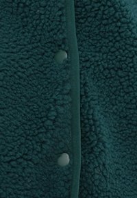 Ύφασμα φλις σε τυρκουάζ με υφή, που διαθέτει λεπτό, πράσινο αντίθετο τελείωμα και ορατό κλείσιμο με κουμπί τύπου τσιμπίδας.