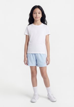 Niña joven de pie, mirando hacia adelante, vistiendo una camiseta blanca, pantalones cortos azul claro, calcetines blancos y zapatillas blancas, sobre un fondo liso.