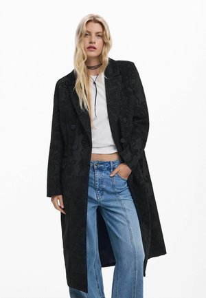 Manteau noir texturé au design long, avec des boutons double boutonnage et des poches latérales, associé à un jean bleu clair ample.