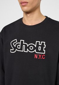 Felpa nera con logo "Schott N.Y.C." in bianco e rosso. Il materiale sembra morbido, con un design a girocollo e una texture minimale.