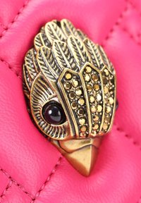 Antieke gouden broche in de vorm van een vogelkop met een zwarte ogen en sierlijke veren op een gequilt, helderroze leren achtergrond.