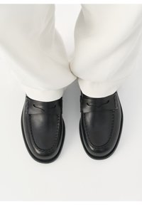 Mocassins penny en cuir noir portés avec un pantalon blanc, vus de dessus sur un fond blanc.