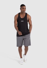Zwarte tanktop met wit logo, gecombineerd met grijze shorts en witte sneakers. De tanktop heeft een klassieke snit en de shorts zijn boven de knie.
