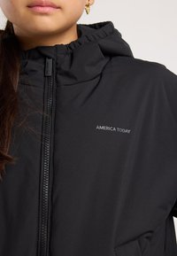 Close-up of person wearing black hooded jacket with "AMERICA TODAY" logo and zipper, showing part of face and gold earring.