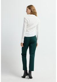 Camicia bianca a maniche lunghe con colletto, abbinata a pantaloni verdi; il modello indossa stivaletti neri con tacco. La vista posteriore mostra una vestibilità sartoriale e polsini.