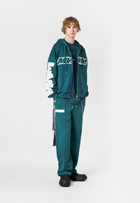 Sudadera color verde azulado con lettering blanco, cierre con cremallera; combinada con pantalones a juego que presentan bolsillos laterales y acentos gráficos; zapatos negros texturizados.