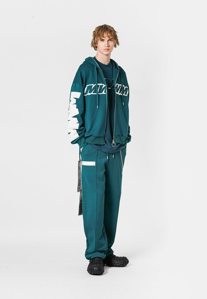 Sudadera color verde azulado con lettering blanco, cierre con cremallera; combinada con pantalones a juego que presentan bolsillos laterales y acentos gráficos; zapatos negros texturizados.