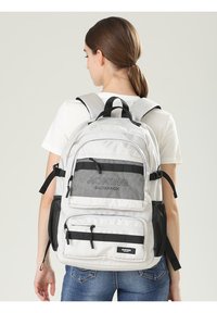 Zaino con tessuto grigio chiaro, compartimenti con doppia cerniera, accenti in rete, spallacci regolabili e maniglia superiore per il trasporto.