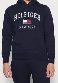 Sudadera navy con bolsillo frontal, con el texto "HILFIGER" y "NEW YORK" en blanco, además de un pequeño logo de bandera. Fabricada con tejido texturizado.