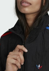 Veste noire à col montant, avec des bandes rouges et bleues sur les épaules et un logo brodé. Tissu lisse et texturé, fermeture éclair.