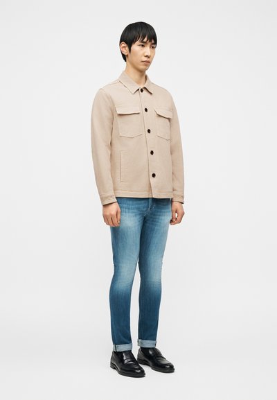 Άνδρας όρθιος, φορά μπεζ σακάκι με κουμπιά, ανοιχτό μπλε ξεβαμμένο τζιν με μανσέτες και μαύρα δερμάτινα παπούτσια, σε απλό λευκό φόντο.