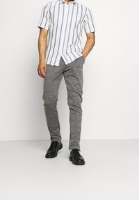 Camicia a maniche corte con base bianca e righe verticali grigie, abbinata a pantaloni a quadri grigi e scarpe in pelle nera.