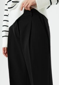 Zwarte geplooide broek gemaakt van gladde stof, met zijzakken, gecombineerd met een witte trui met zwarte strepen.