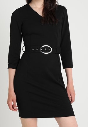 Vrouw die een nauwsluitende zwarte jurk draagt met driekwart mouwen en een brede zwarte riem met een grote zilveren ovale gesp.