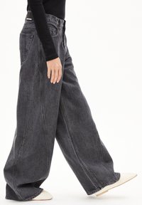 Schwarze Denim-Jeans mit weitem Bein und hoher Taille, die subtilen Verfärbungen und Stitching-Details aufweisen, kombiniert mit einem hellen Schuh.