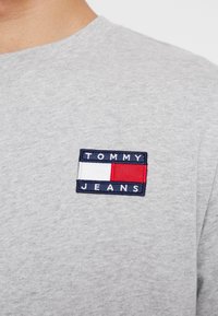 Graues Baumwoll-T-Shirt mit einem gestickten Patch in Marineblau, Rot und Weiß mit dem Text "TOMMY JEANS". Klassisches Rundhalsdesign.