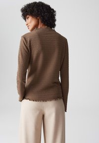 Donna con capelli ricci che indossa una maglietta lunga marrone a trama e pantaloni beige, di spalle contro uno sfondo grigio chiaro uniforme.