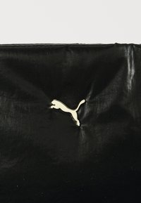 Crna torba s sjajnom, teksturiranom površinom koja na strani ima zlatni logo skakavca pumeg. Glatki materijal i minimalistički dizajn.
