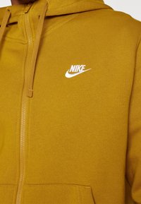 Sudadera con cremallera amarilla hecha de tejido suave, con un bolsillo tipo canguro y el logo de Nike bordado en blanco en el lado izquierdo del pecho.