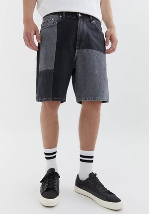 Homme portant un short en jean patchwork noir et gris, des chaussettes blanches à rayures noires et des baskets noires à semelles blanches.