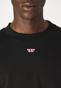 Črna majica s kapuco z ribanim izrezom in majhnim rdečim vezenim logotipom na prsih. Gladka tekstura, sproščen oblikovanje.